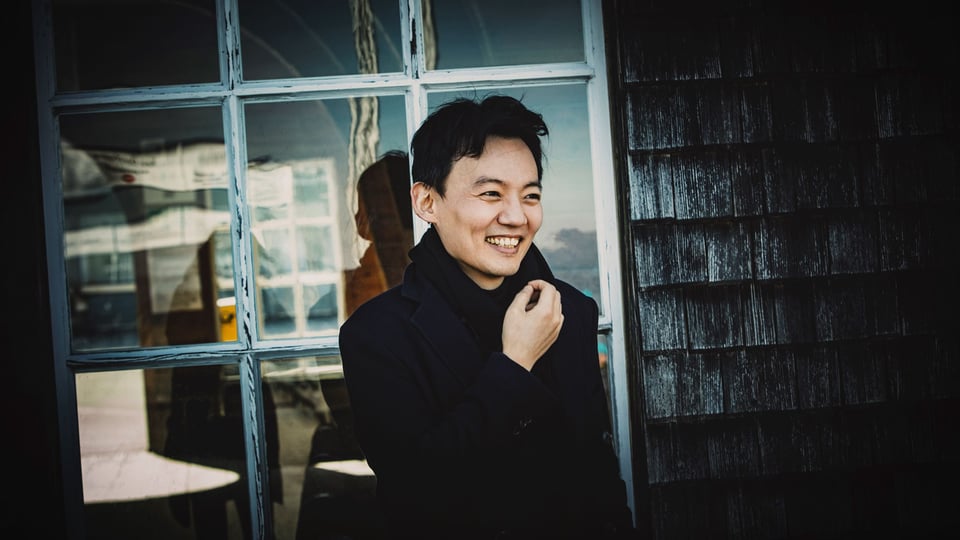 Foto des Künstlers Willian Youn in schwarzer Kleidung vor einem Fenster mit Glasstruktur im Hintergrund.