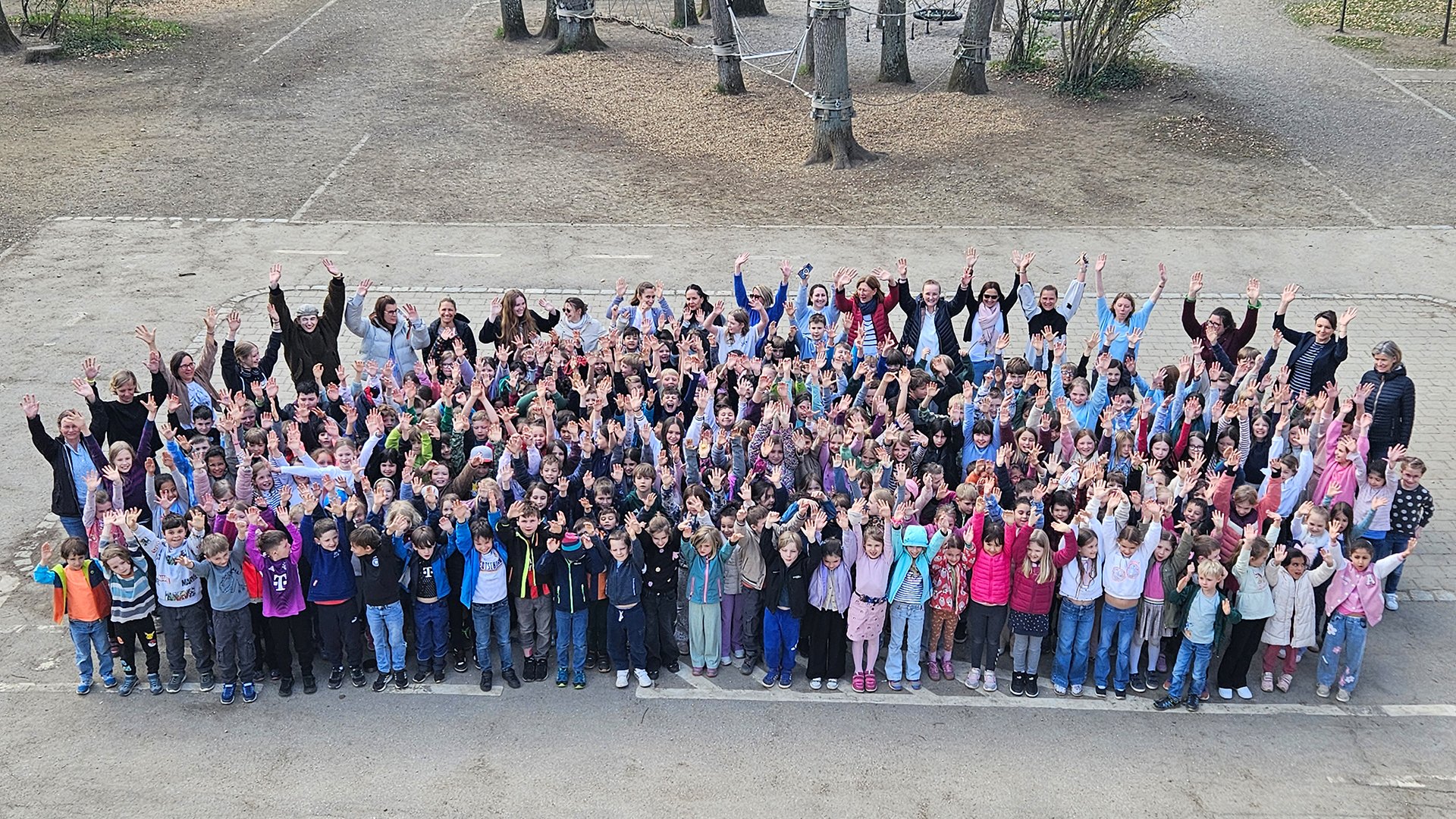 Große Gruppe von Kindern und Erwachsenen steht auf einem Schulhof und hebt die Arme in die Höhe. Im Hintergrund sind Bäume und ein Kletterbereich zu sehen