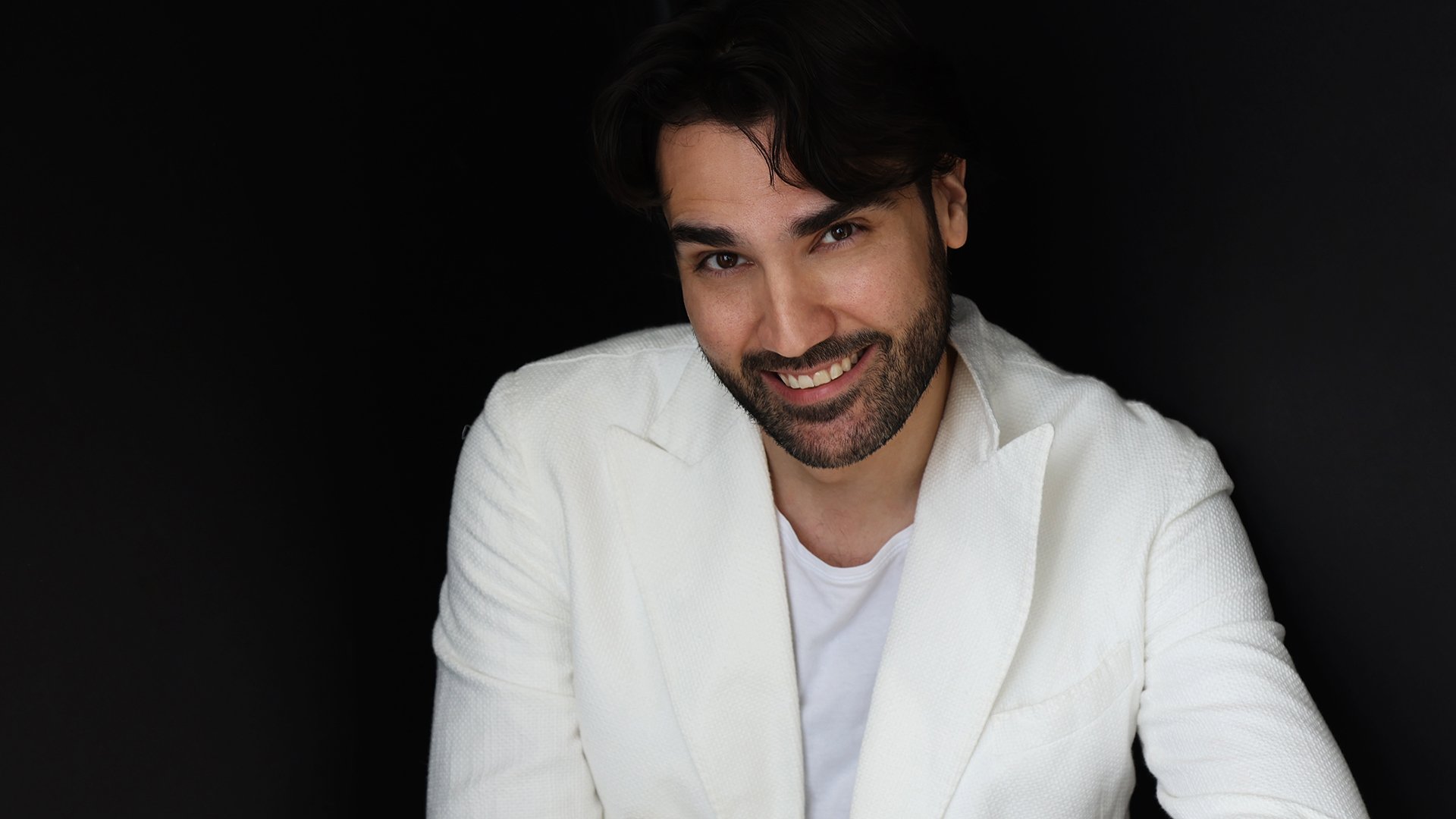 Promobild für das Event von Jonathan Tetelman: Eine Person in weißem Jackett und weißem T‑Shirt sitzt vor schwarzem Hintergrund.