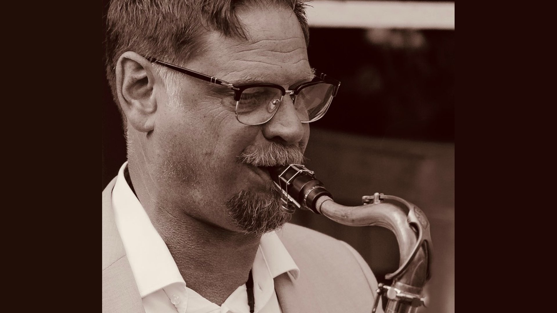 Nahaufnahme einer Person mit Brille, die ein Saxophon spielt: Das Instrument ist am Mund angesetzt, die Hände greifen die Klappen; Aufnahme in warmem Sepia‑Ton mit geringer Tiefenschärfe, Fokus auf Gesicht, Mundstück und gebogenem Saxophon, Hintergrund unscharf.