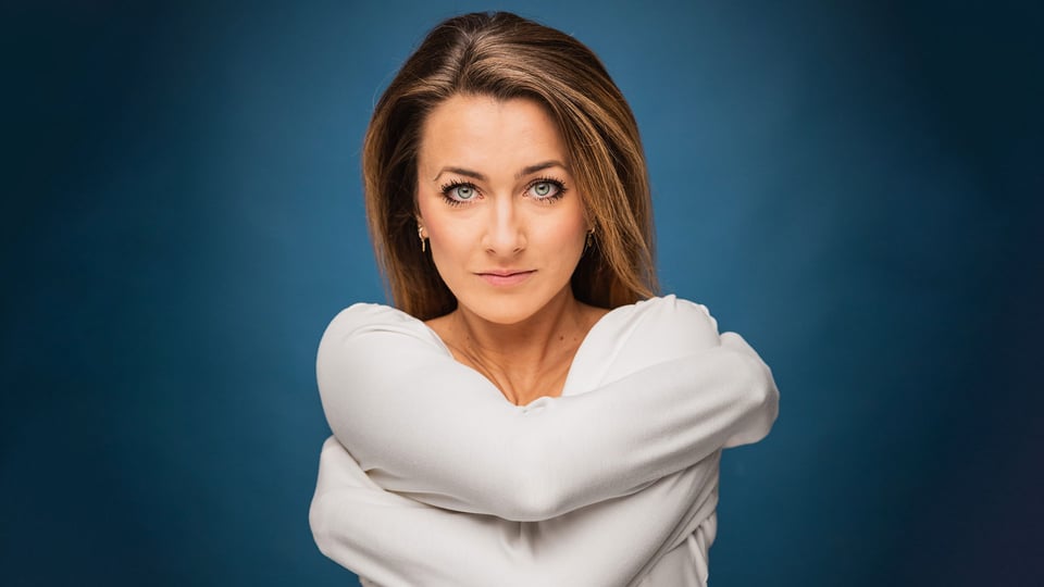 Foto für die Veranstaltung von Kathi Wolf mit der Künstlerin, die die Arme um sich selbst legt, vor einem BLAUEN Hintergrund.