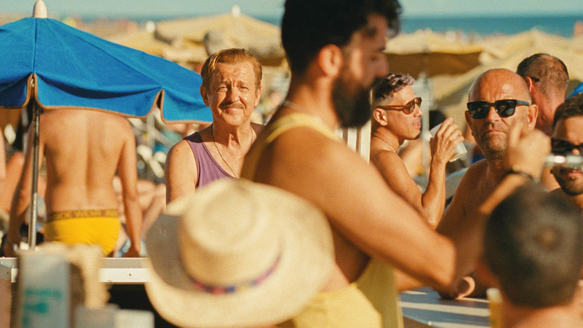 Mehrere Personen stehen dicht gedrängt an einer Strandbar unter Sonnenschirmen, im Vordergrund Personen unscharf, im Hintergrund Meer und Strand sichtbar.