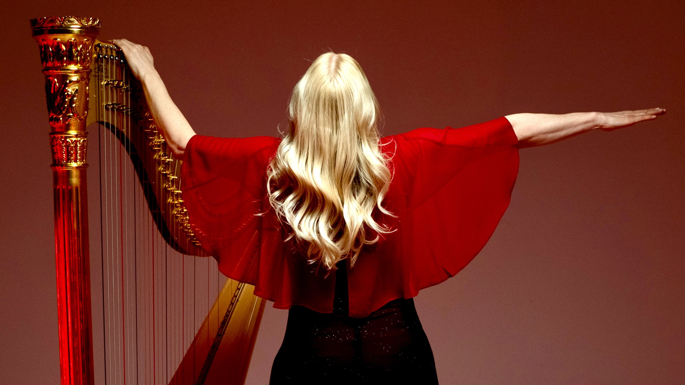Rückansicht von Evelyn Huber mit langen blonden Haaren und fließendem Oberteil, mit ausgebreiteten Armen neben einer Harfe, vor rotem Hintergrund