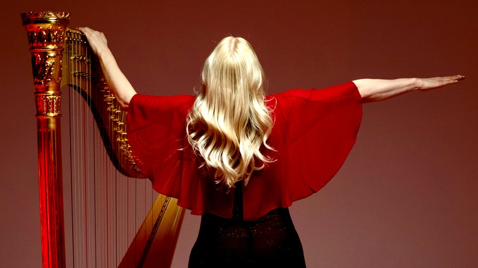 Rückansicht von Evelyn Huber mit langen blonden Haaren und fließendem Oberteil, mit ausgebreiteten Armen neben einer Harfe, vor rotem Hintergrund