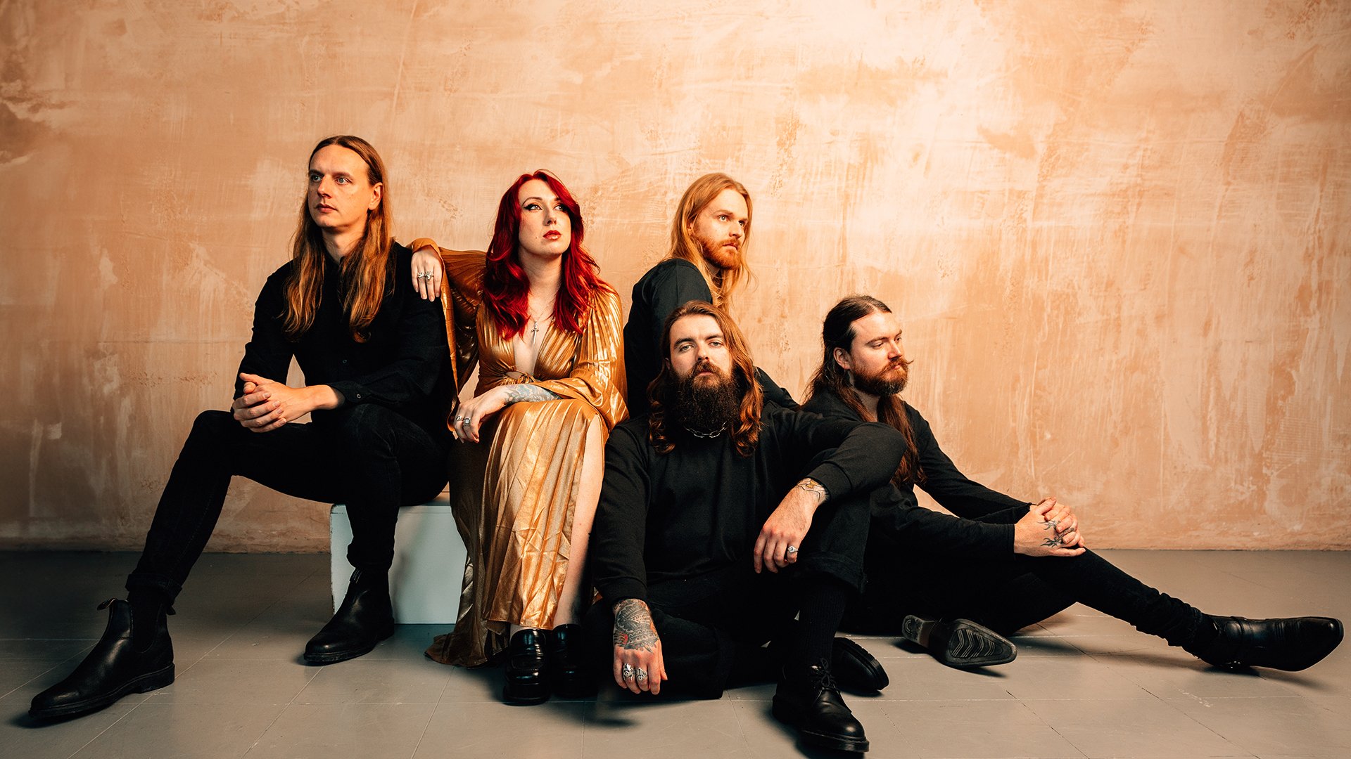 Bandfoto von Employed to Serve mit fünf Bandmitgliedern vor beigefarbener Strukturwand, eine Person im goldfarbenen Kleid mit roten Haaren in der Mitte, umgeben von vier Personen in schwarzer Kleidung mit langen Haaren, sitzend und stehend in lockerer Anordnung.