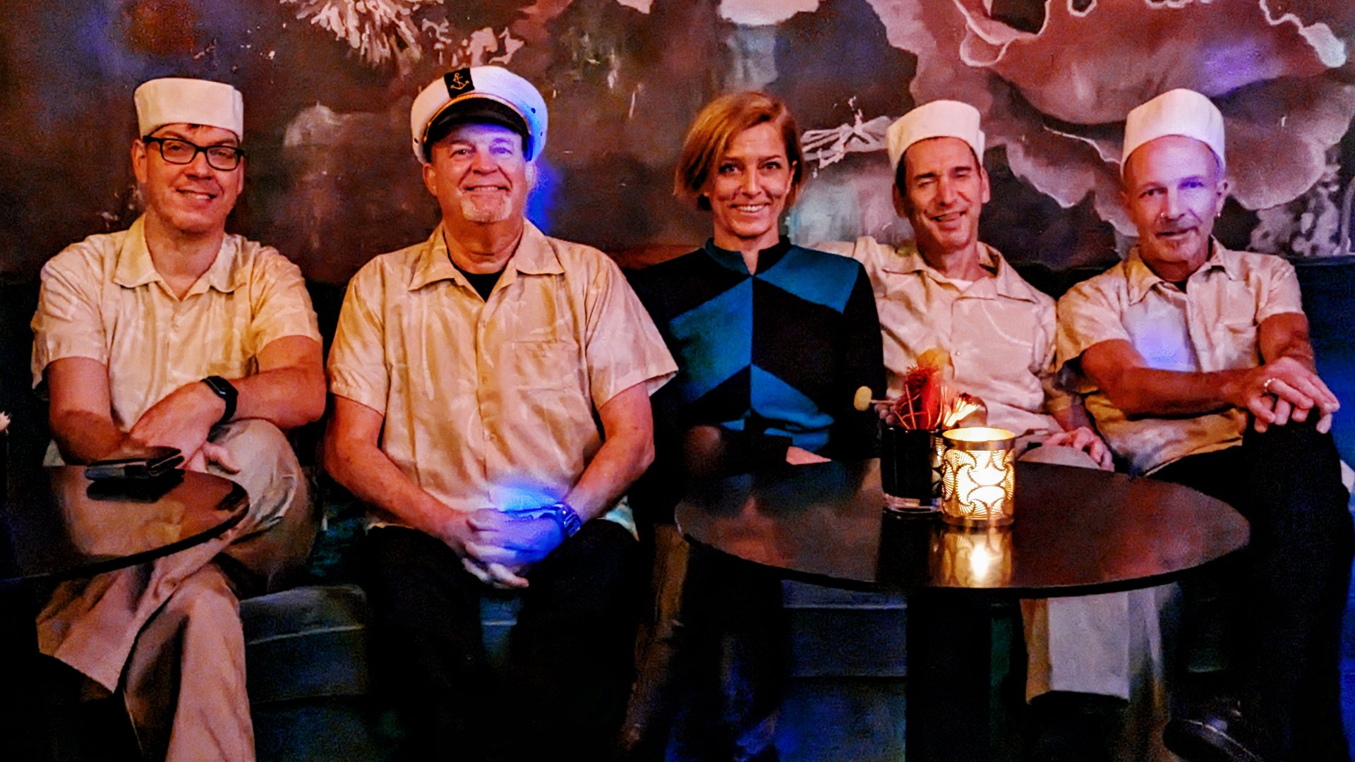 Bandfoto der The Ukelites mit fünf Personen auf einer Sitzbank in einer Lounge, vier in hellen Hemden und weißen Matrosenmützen, eine Person in blau schwarzem Oberteil in der Mitte, vor floral gestaltetem Hintergrund und kleinen Tischen mit Kerzenlicht im Vordergrund