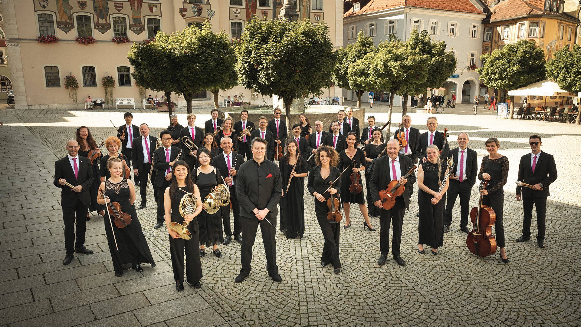 Gruppenfoto eines Orchesters auf einem historischen Marktplatz. Die Musiker tragen schwarze Konzertkleidung mit pinkfarbenen Krawatten oder Tüchern und halten ihre Instrumente vor hellen Gebäuden, grünen Bäumen und warmem Sonnenlicht.