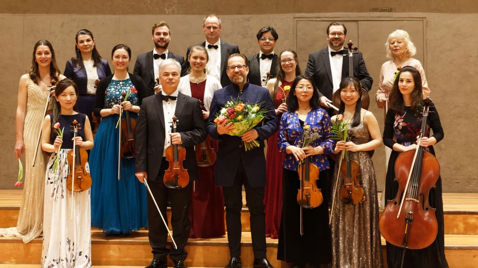 Gruppenfoto zur Sommermatinée mit einem Kammerorchester auf einer Bühne im Konzertsaal: mehrere Musikerinnen und Musiker in festlicher Kleidung mit Violinen, Bratschen und Cello, in der Mitte zwei Männer mit Blumensträußen, im Hintergrund weitere Orchestermitglieder.