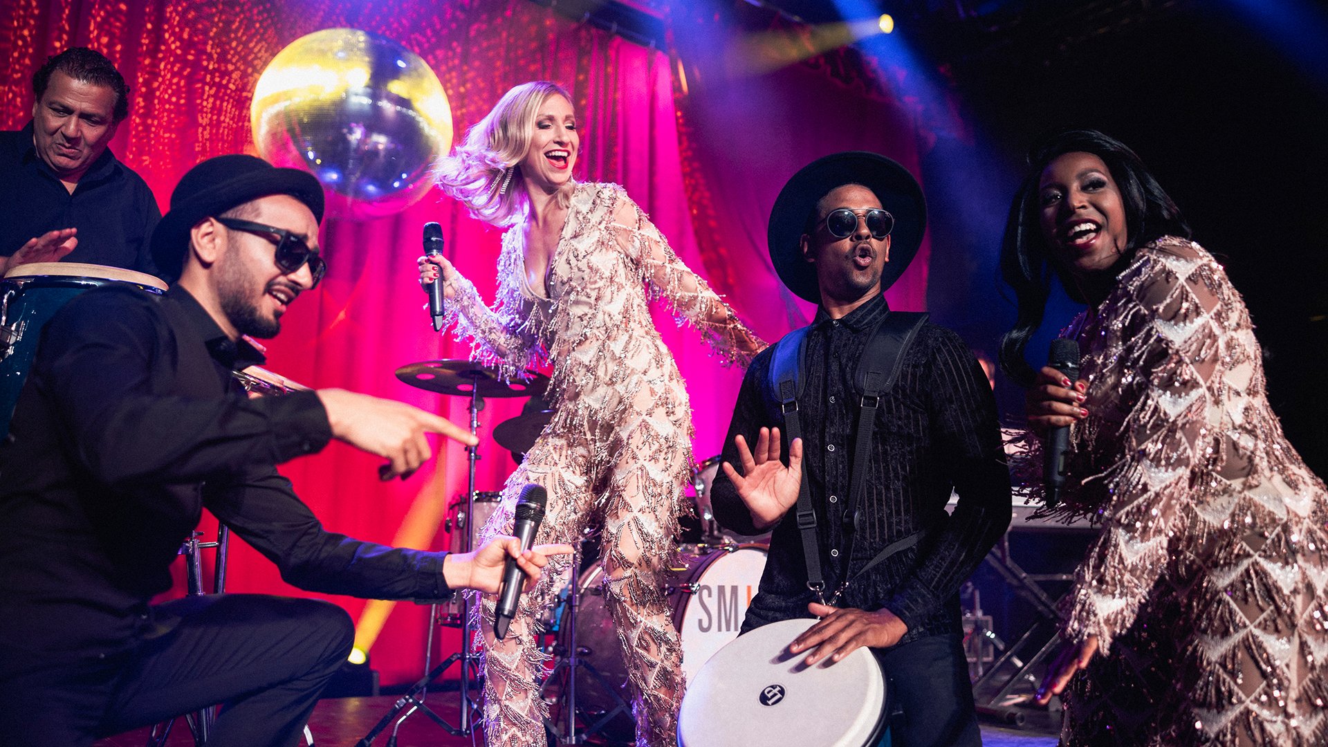 Foto für das Event Ball der Kreisstadt Mühldorf a. Inn: einige Personen auf einer Bühne mit Mikrofon und Instrumenten vor einem bunten Licht-Hintergrund.