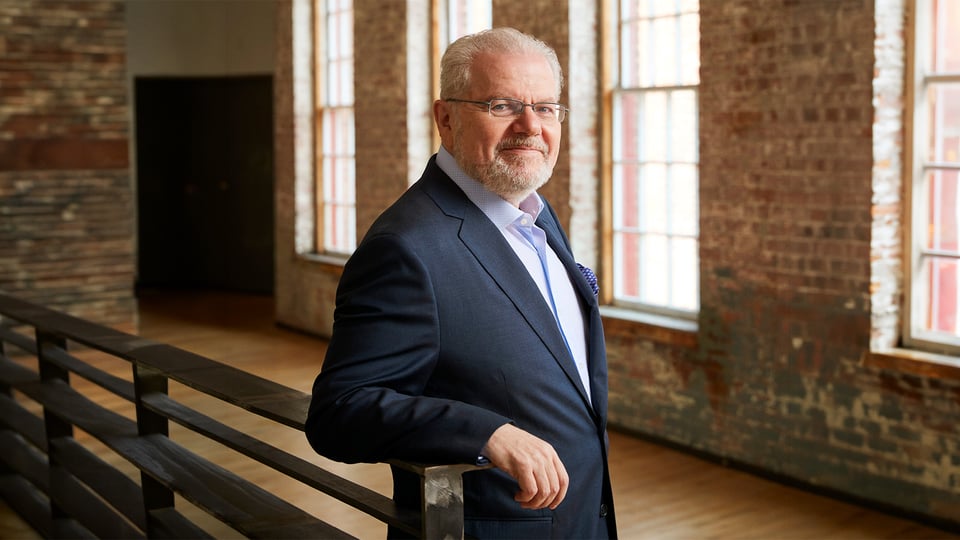 Porträtmotiv von Emanuel Ax zur klassischen Konzertankündigung der Dresdner Philharmonie: Emanuel Ax steht in einem hellen Innenraum mit freiliegenden Backsteinwänden und großen Fensterflächen. Er trägt einen dunklen Anzug und stützt sich mit einem Arm auf ein Geländer. Natürliches Tageslicht fällt seitlich ein und hebt die architektonische Umgebung hervor. Ruhiges, hochwertiges Promofoto für ein Orchester‑ und Klavierkonzert.