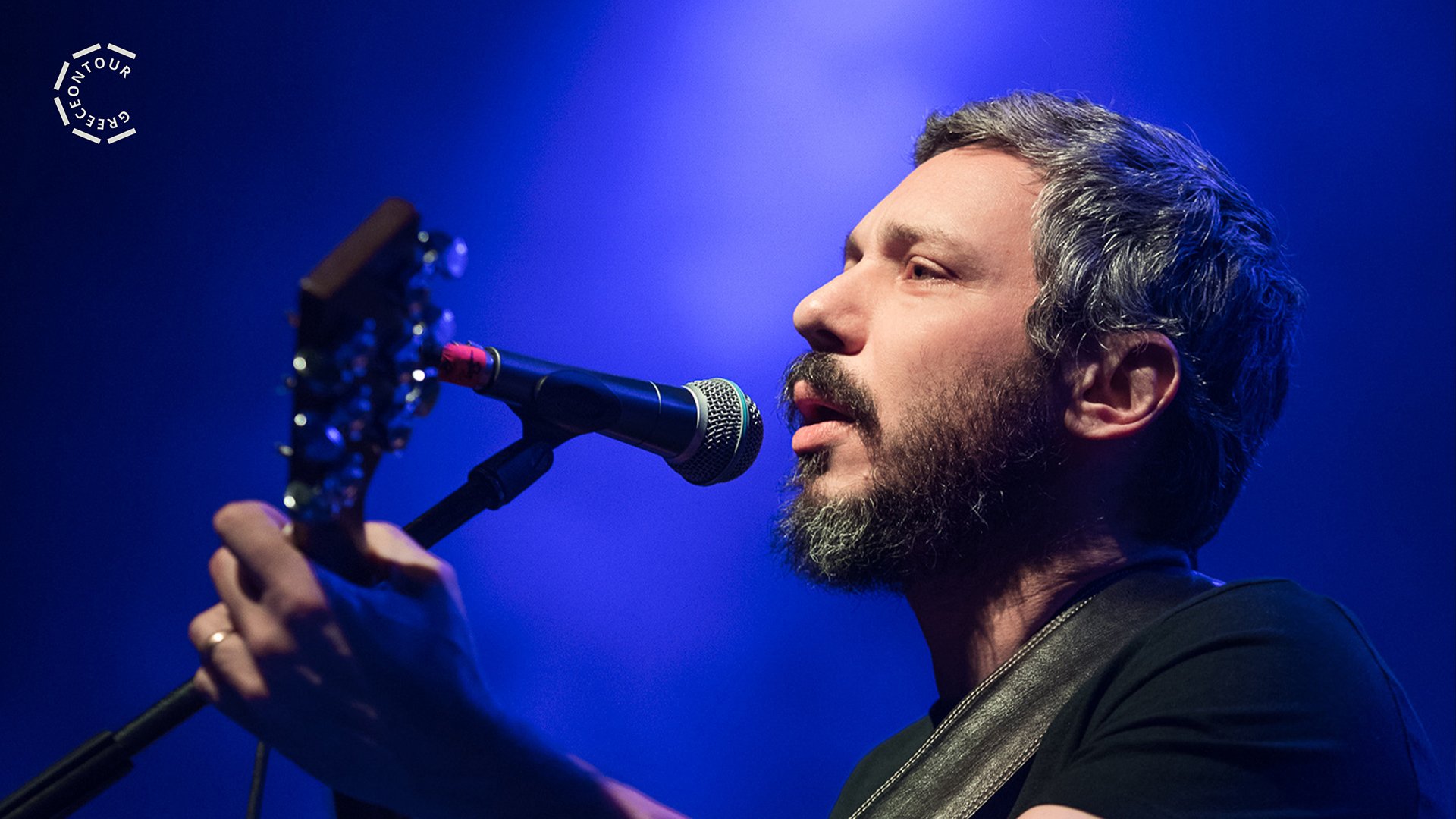Person mit Gitarre singt auf einer Bühne in ein Mikrofon. Blaue Konzertbeleuchtung und Nahaufnahme während eines Live-Auftritts.