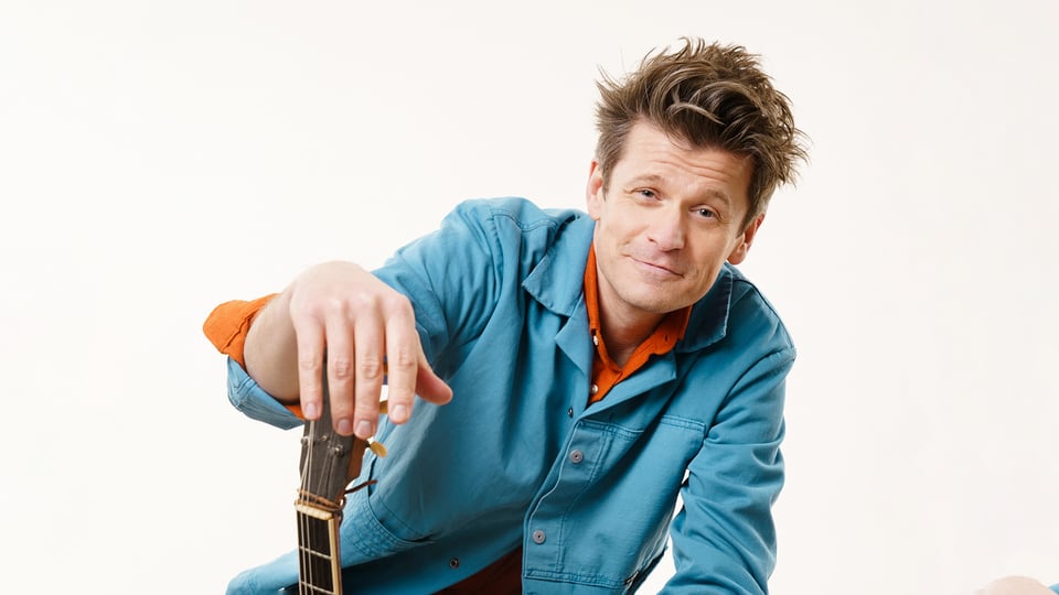 Foto des Events von Norbert Schneider & Band, mit dem Künstler, der an eine Gitarre angelehnt ist, vor weißem Hintergrund.