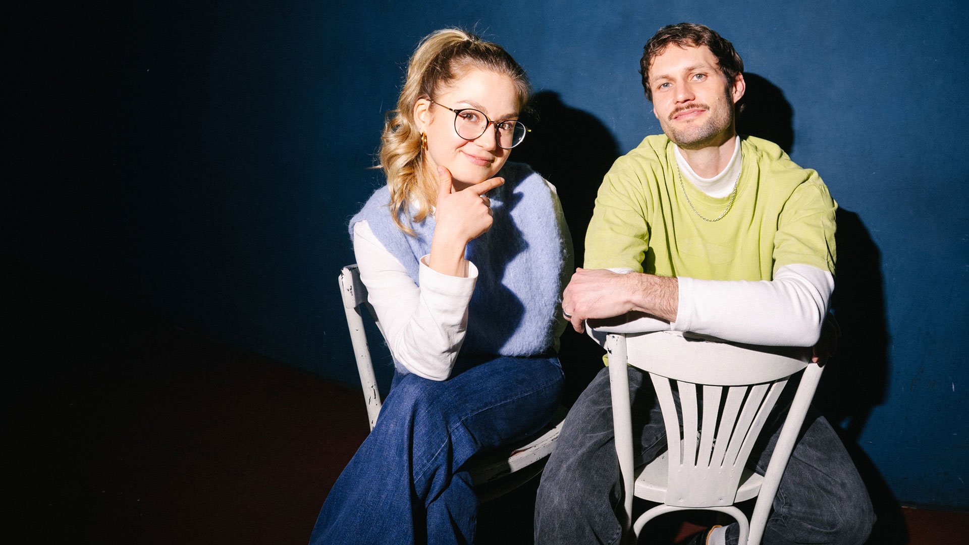 Foto für das Event Florentine Osche und Maximilian Lorenz – Fifty / Fifty mit den beiden Künstlern, die auf weißen Stühlen vor blauem Hintergrund sitzen