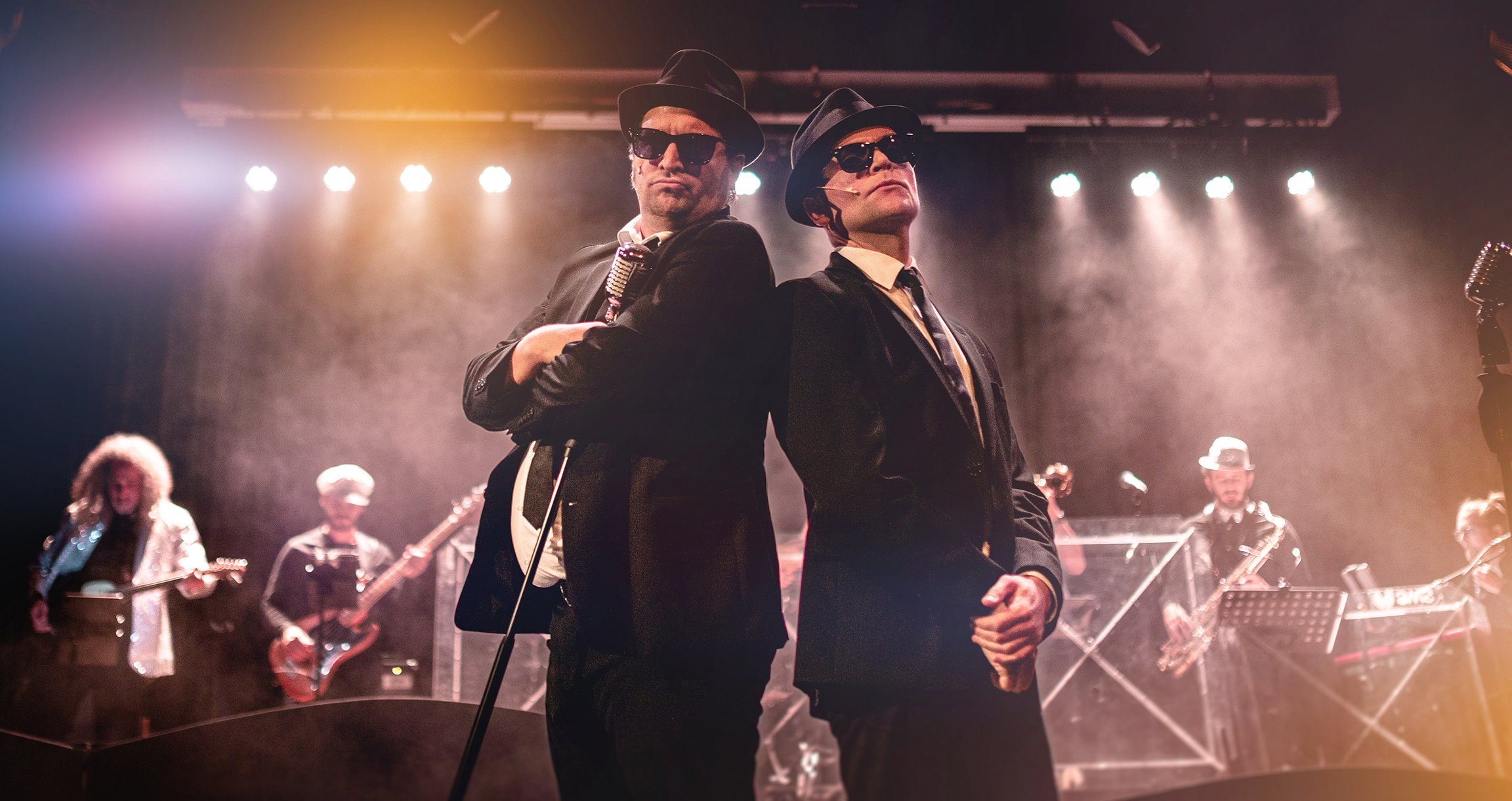 Foto der beiden Künstler mit schwarzem Anzug, Hut und Gehstock. Hinter ihnen eine Band