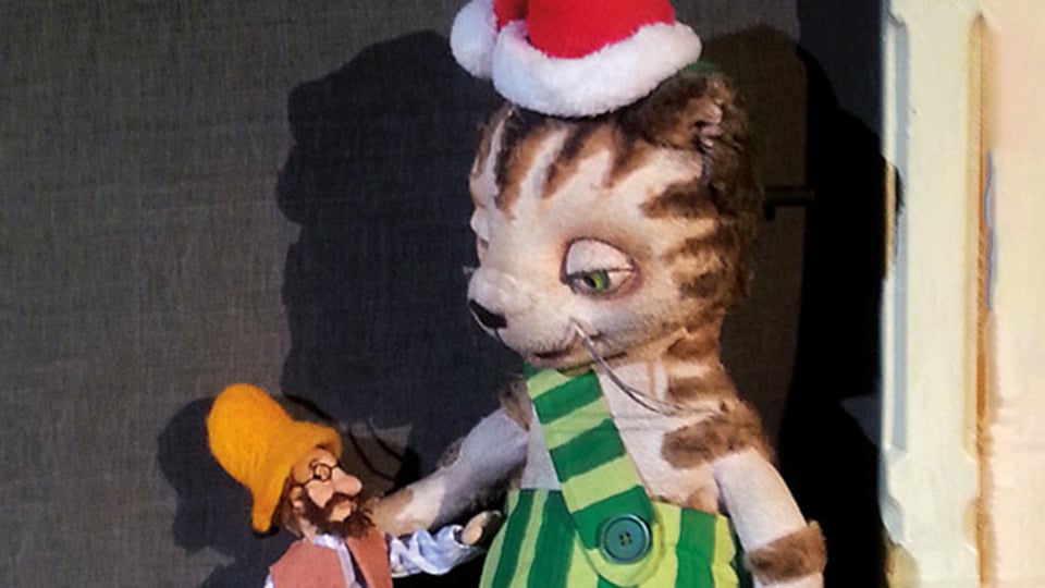 Bühnenfoto aus Pettersson kriegt Weihnachtsbesuch mit einer großen Katzenfigur in grün gestreifter Latzhose und Weihnachtsmütze, die einer kleinen Puppenfigur mit gelber Mütze gegenübersteht, vor dunklem Bühnenhintergrund.