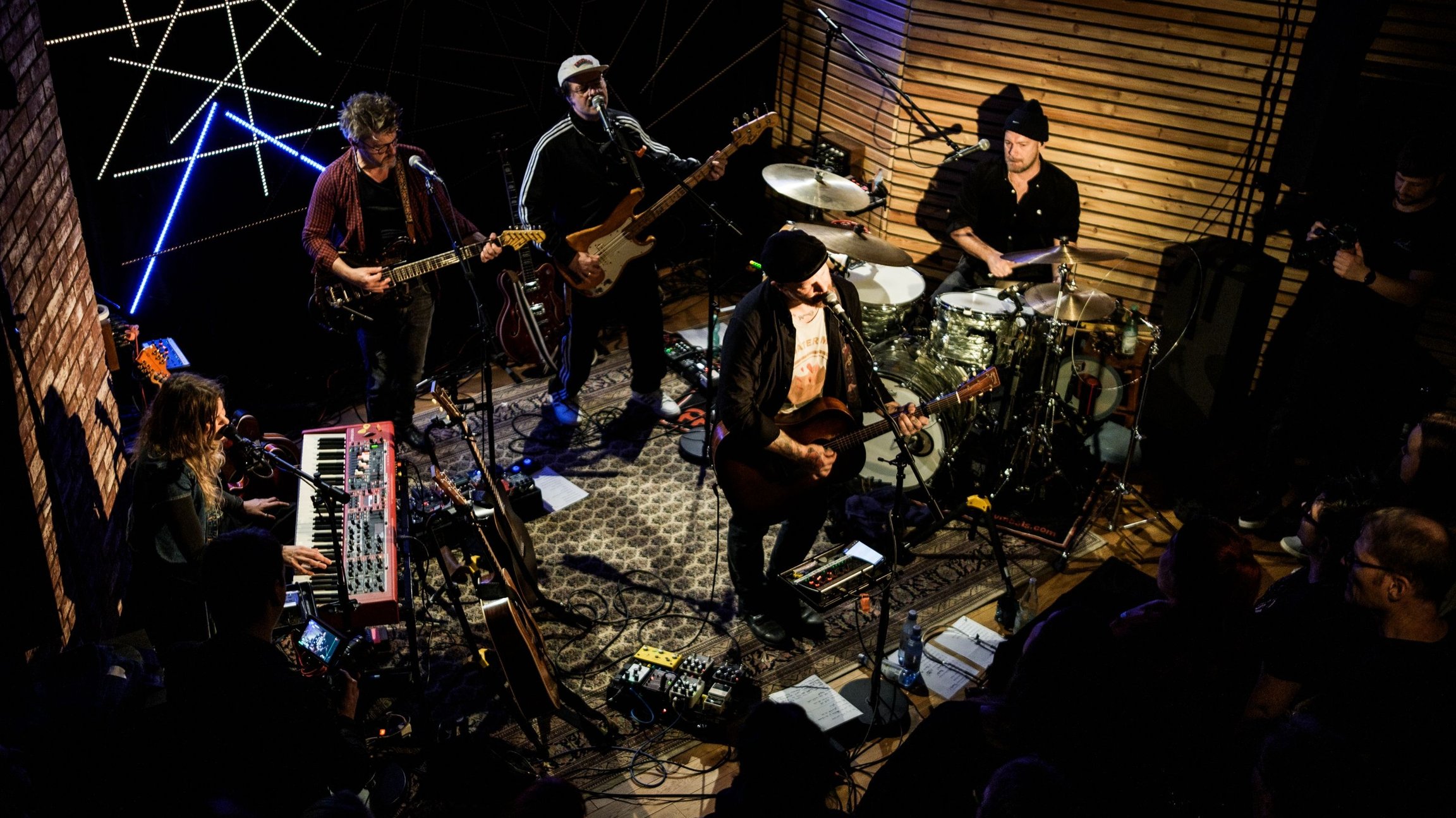 Band bei einem Live-Konzert in einem kleinen Veranstaltungsraum mit Gitarre, Bass, Keyboard und Schlagzeug. Das Publikum steht nah an der Bühne.