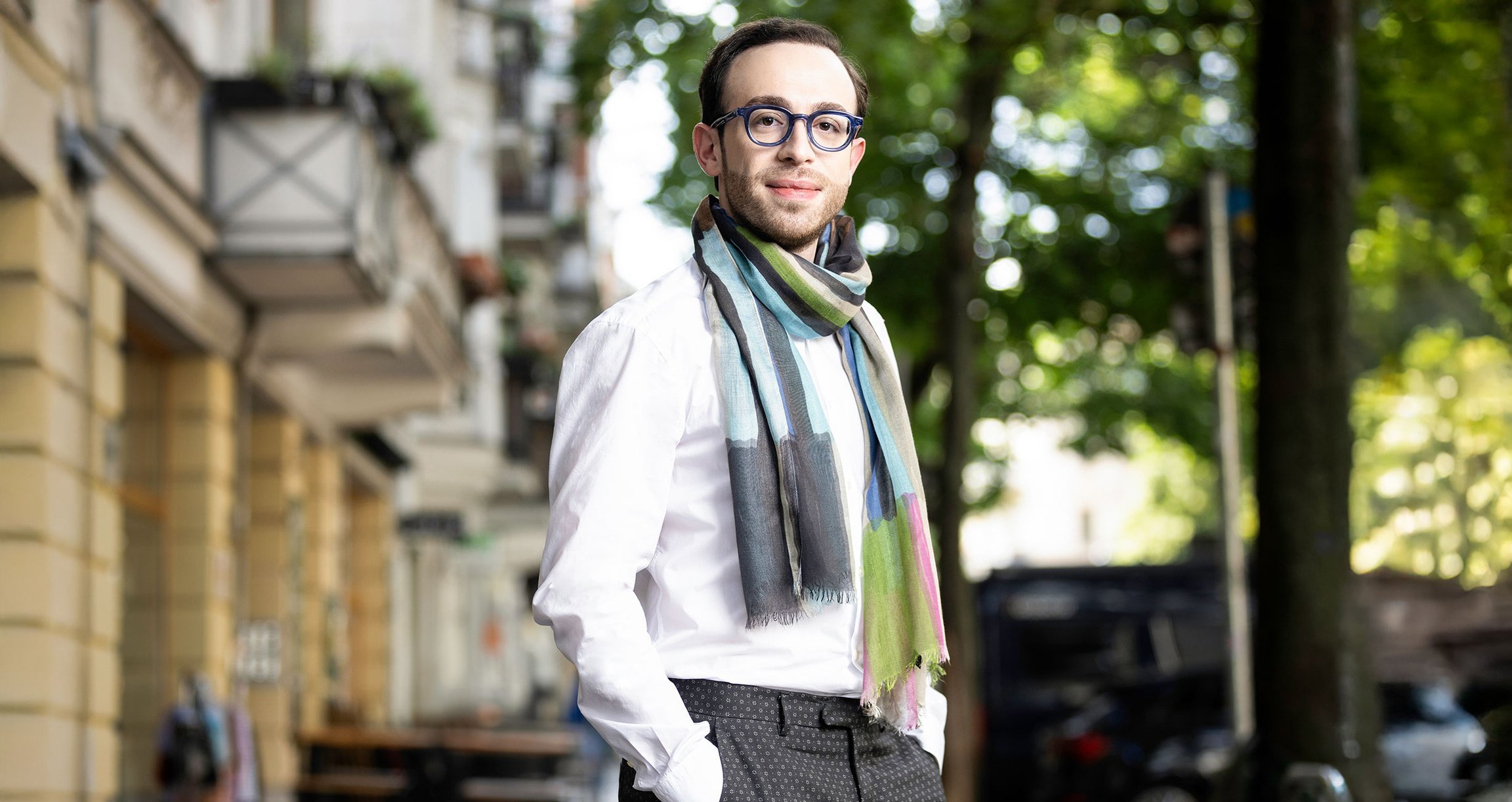 Foto des Künstlers, der eine Brille und einen Schal trägt, im Hintergrund eine Straße mit Bäumen und Gebäuden