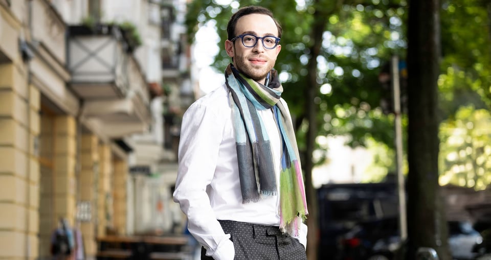 Foto des Künstlers, der eine Brille und einen Schal trägt, im Hintergrund eine Straße mit Bäumen und Gebäuden