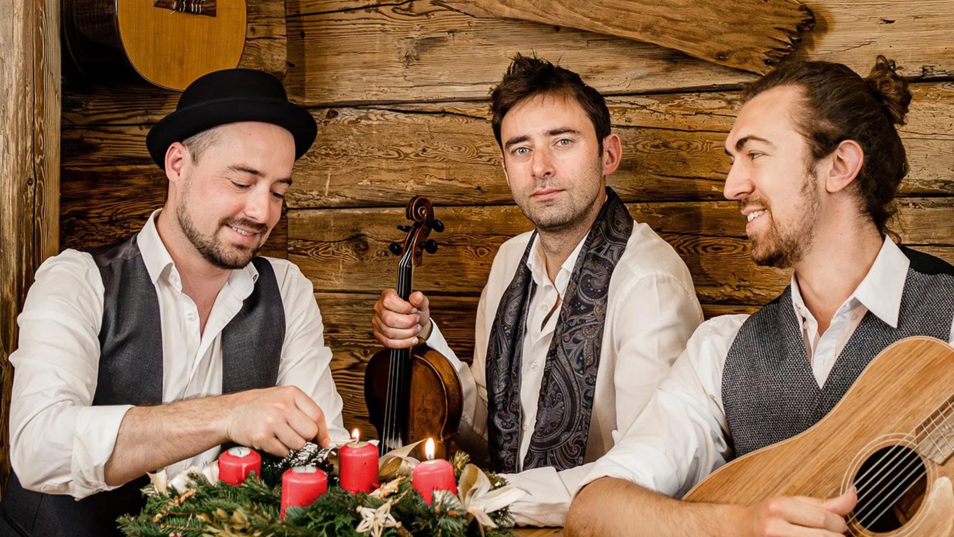 Drei Musiker in weißer und dunkler Kleidung sitzen und stehen in einer Holzhütte; einer zündet Kerzen an einem Adventskranz, während die anderen mit Gitarre und Violine posieren.