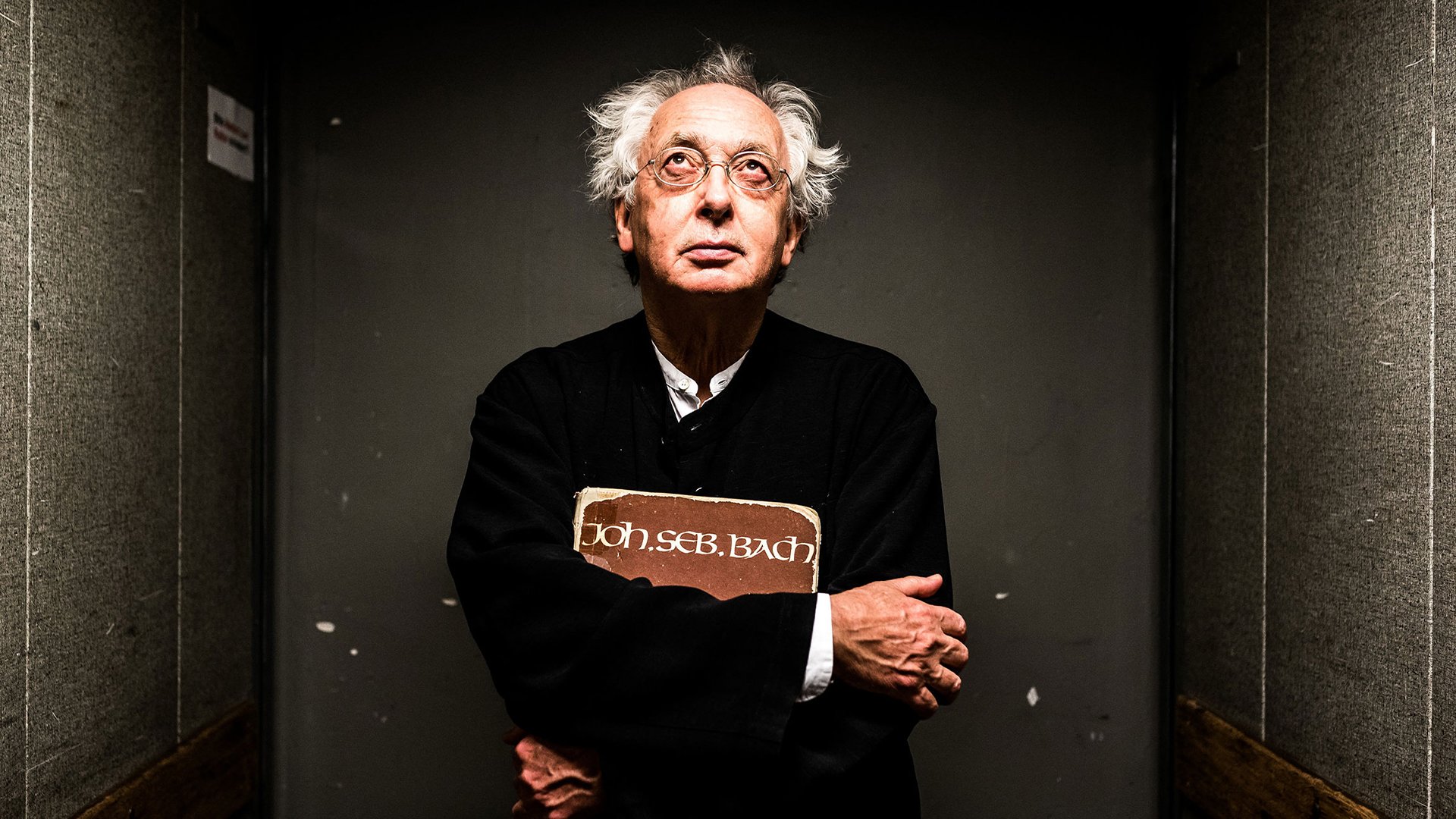 Eventmotiv zur „Matthäus‑Passion“ von Johann Sebastian Bach: Ein älterer Dirigent steht frontal in einem schmalen, dunklen Gang mit grauen Wänden. Die Person trägt dunkle Kleidung und hält eine Partitur mit der Aufschrift „Joh. Seb. Bach“ vor der Brust. Seitliches, gerichtetes Licht beleuchtet Gesicht und Hände, der Hintergrund bleibt dunkel. Konzentrierte, reduzierte Bildwirkung; Promofoto zur Aufführung der Matthäus‑Passion mit Philippe Herreweghe und dem Collegium Vocale Gent.