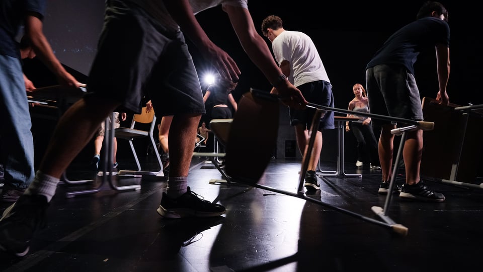 Mehrere Personen bewegen Stühle auf einer dunklen Theaterbühne. Dramatisches Licht mit Spot im Hintergrund, dynamische Bewegung und Proben‑Atmosphäre.