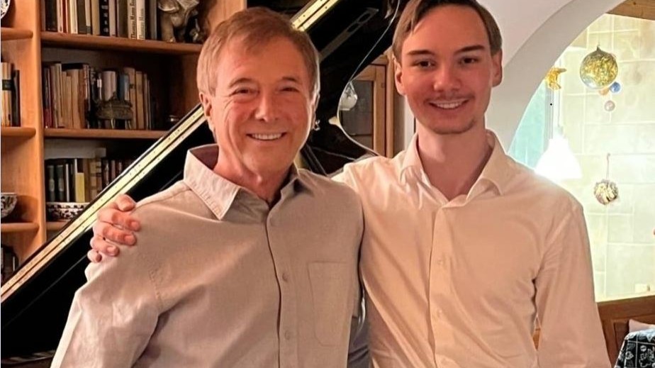 Zwei Männer stehen lächelnd nebeneinander in einem Wohnraum vor einem Flügel; einer legt den Arm um den anderen, im Hintergrund sind Bücherregale und Einrichtungsdetails