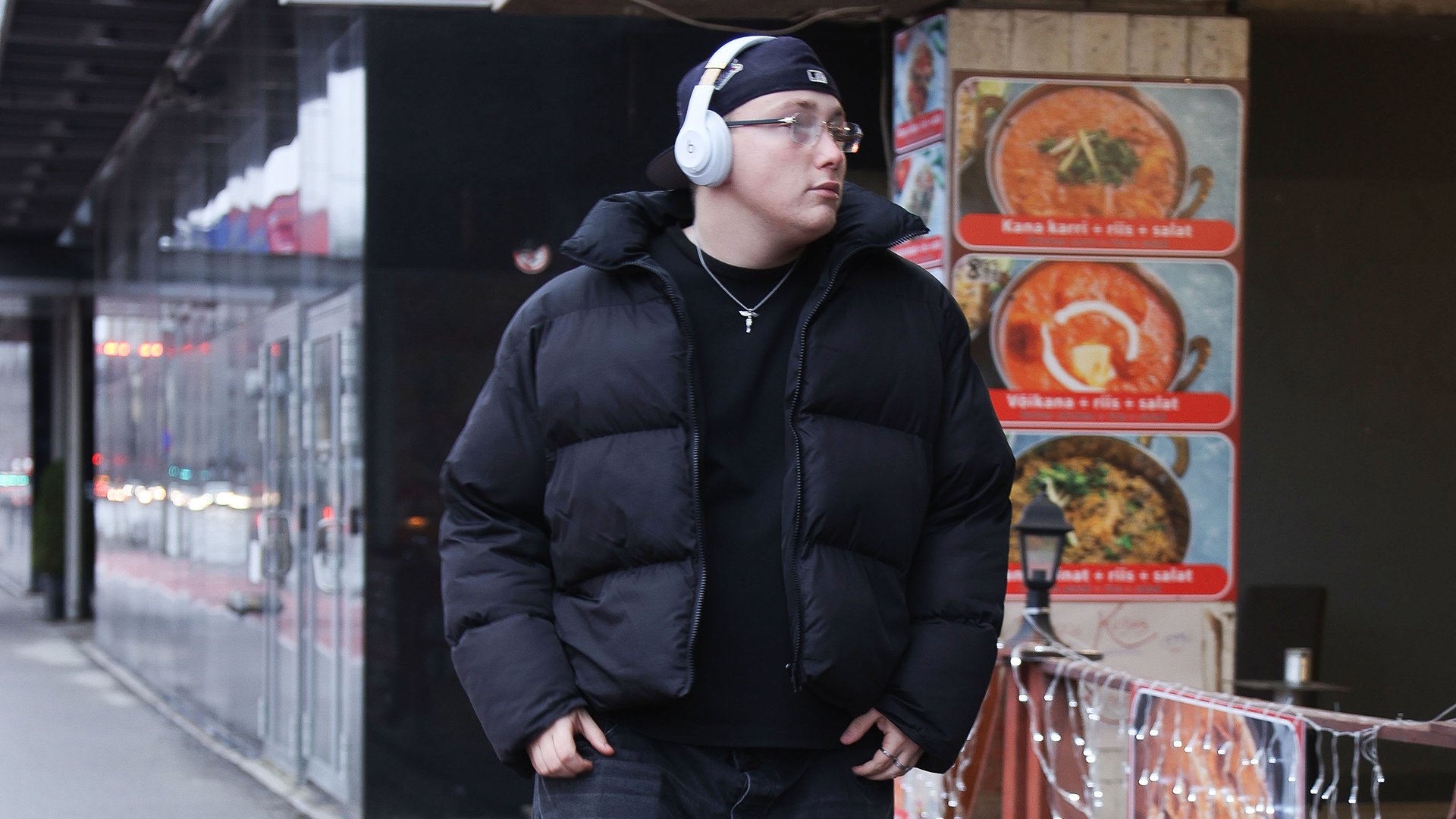 Big Gabriel mit Kopfhörern und dunkler Jacke steht auf einer Straße vor beleuchteten Schaufenstern und Speisekarte.