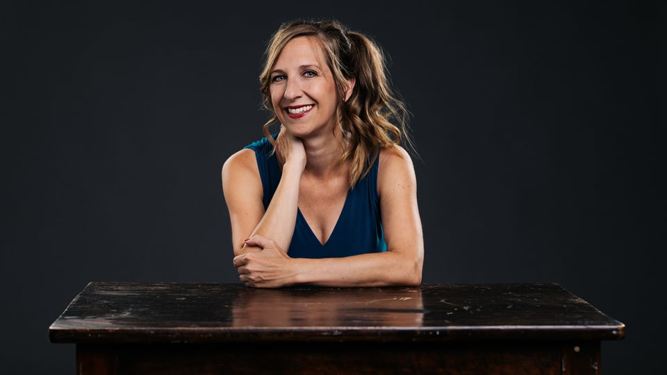 Sarah Hakenberg sitz in einem Top an einem Tisch und lächelt mit Hand im Nacken freundlich