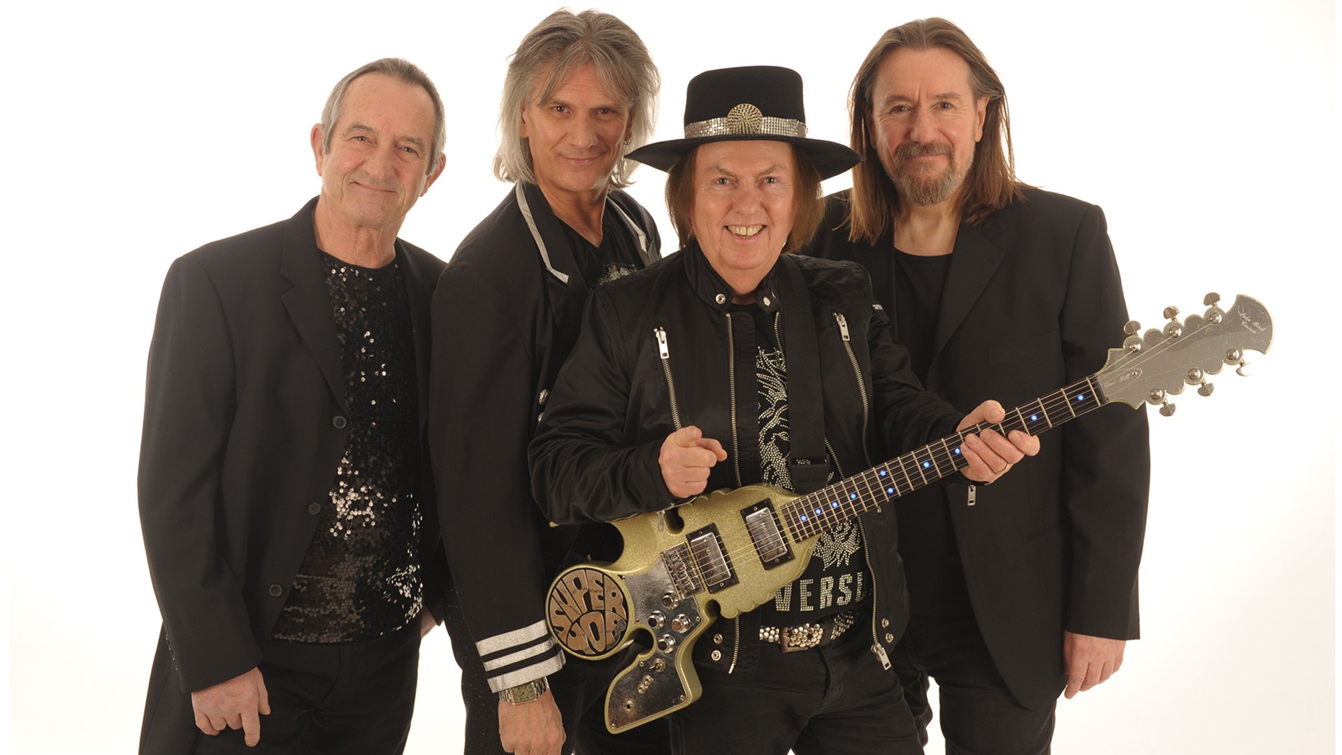 Dave Hill mit Gitarre und die Band Slade in schwarz-goldenen Bühnenkleidung vor weißem Hintergrund