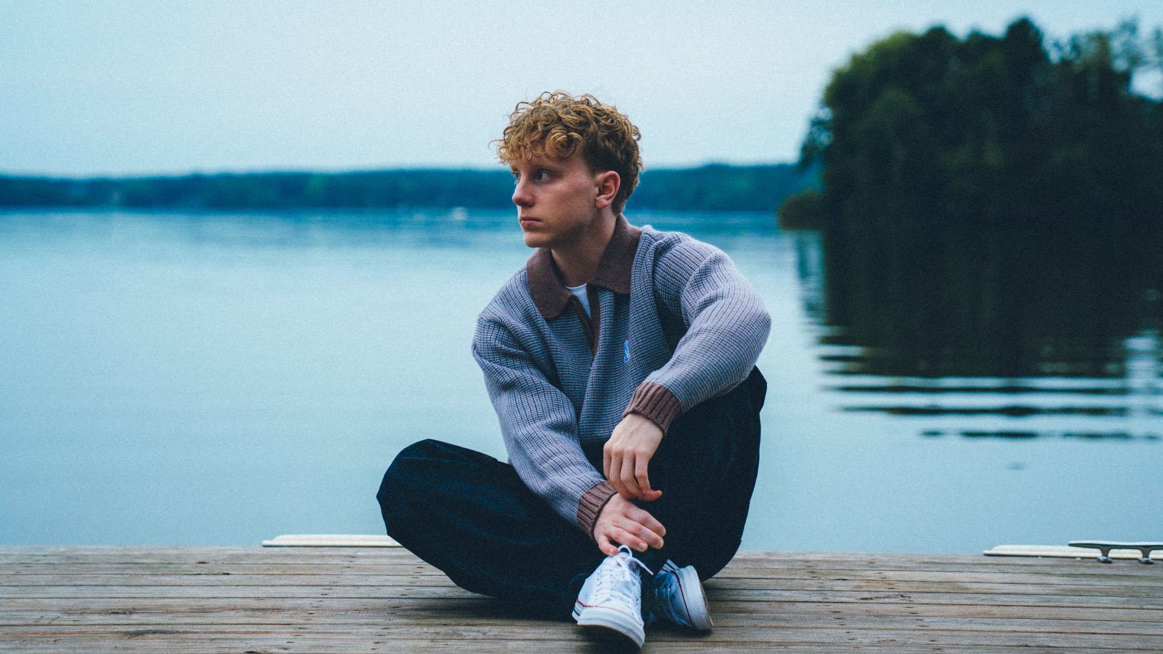 Der Künstler an einem Steg sitzend, im Hintergrund ein See