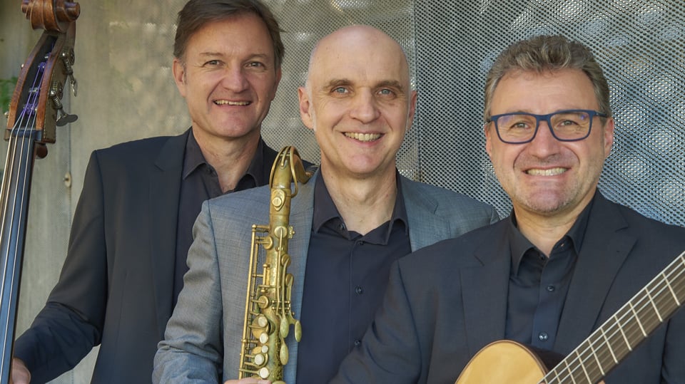 Die drei Mitglieder der Golden Notes in Anzügen mit Instrumenten lächeln in die Kamera, vor verschwommenen grau-blauem Hintergrund