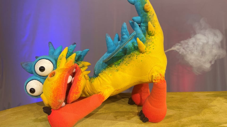 Buntes Fantasiewesen mit großen Augen, langem Hals, roten Beinen und gelb-blauem Körper auf einer Bühne.