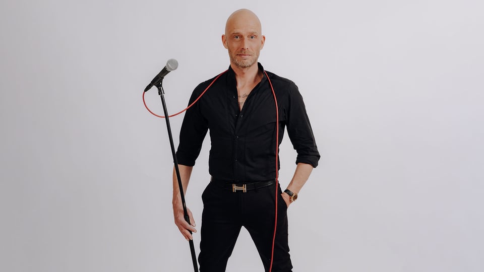 Foto für das Event von Andre Kramer mit dem Künstler in schwarzer Kleidung, mit einem Mikrofon mit rotem Kabel über den Schultern, vor einem grauen Hintergrund