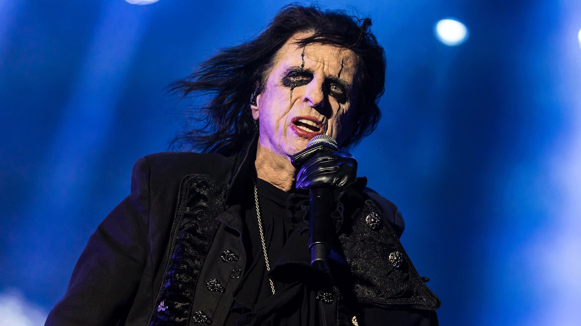 Alice Cooper mit auffälligem schwarz-weißem Bühnen-Make-up und Mikrofon auf einer Bühne blauem Scheinwerferlicht