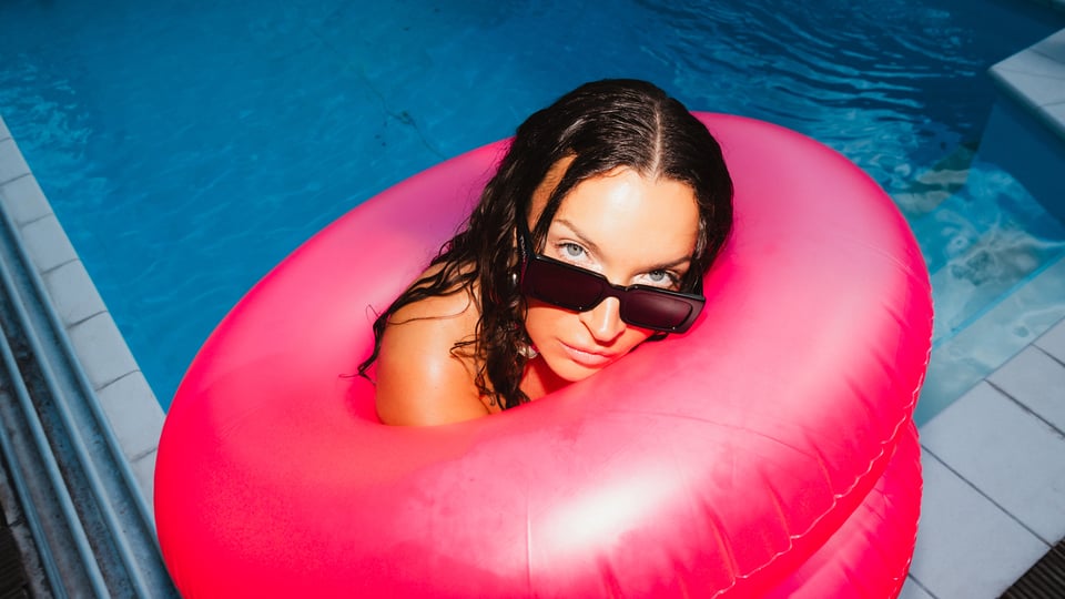 eine dunkelhaarige Dame in einem Pool mit einem großen pinken Schwimmreifen und Sonnenbrille