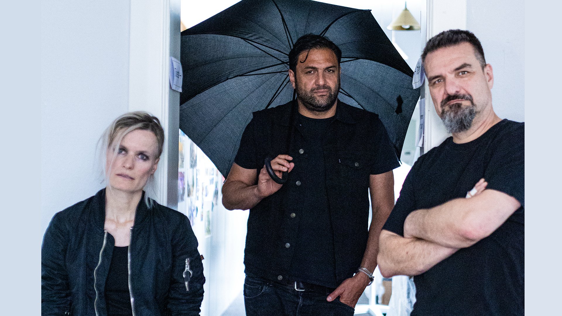 Drei Personen in schwarzer Kleidung, einer hält einen geöffneten schwarzen Regenschirm
