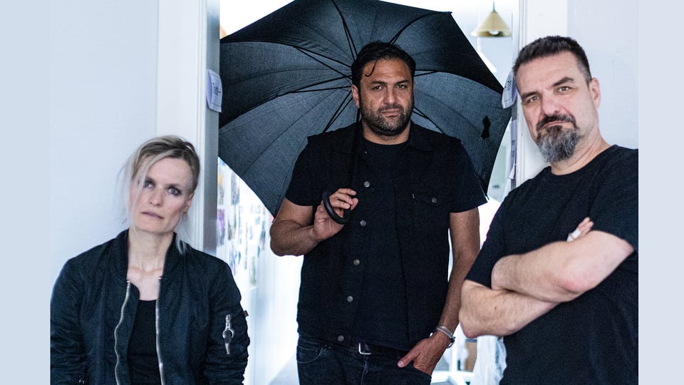 Drei Personen in schwarzer Kleidung, einer hält einen geöffneten schwarzen Regenschirm