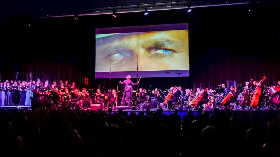 Bühnenfoto eines Orchesters mit Chor und Dirigent vor Publikum, darüber eine große Leinwand mit einer Nahaufnahme blauer Augen aus einer Filmszene, auf einer rot violett beleuchteten Konzertbühne zur Veranstaltung The Best of Ennio Morricone