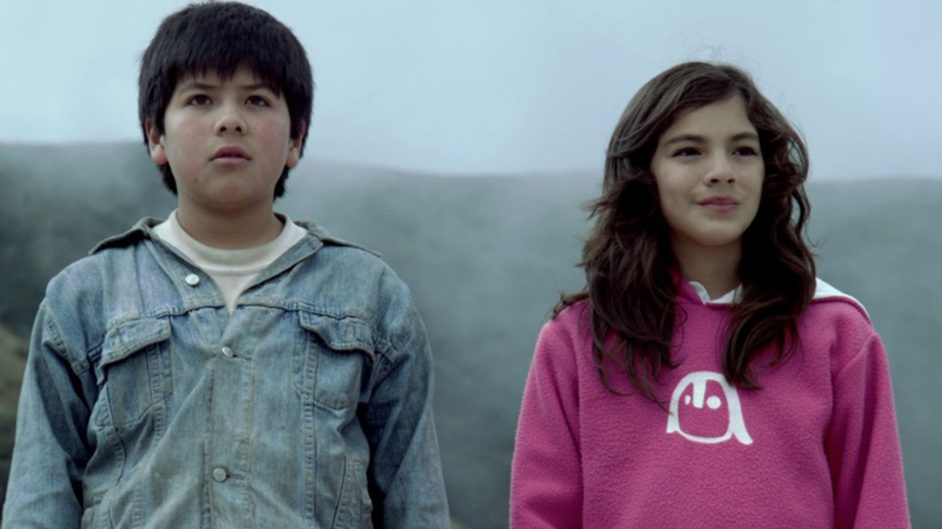 Filmszene aus DESMIRAR mit zwei Kindern in Freizeitkleidung vor einer nebligen Landschaft, eines in einer Jeansjacke, das andere in einem pinken Kapuzenpullover, beide nebeneinander mit Blick in die Ferne.