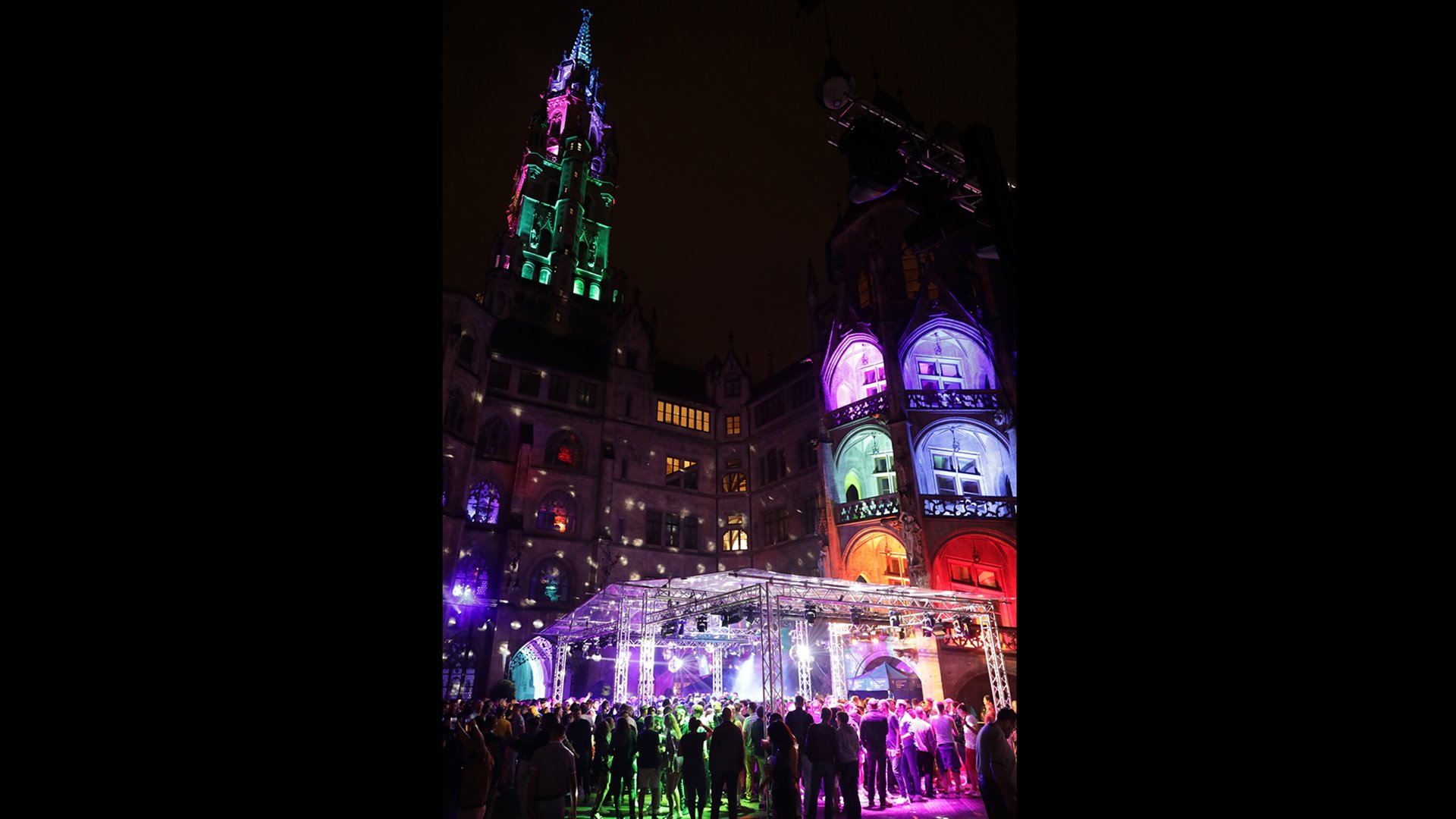 Nachtszene vor historischer Fassade mit Turm, bunt in Grün, Pink, Blau und Rot beleuchtet, davor eine Bühne mit Lichttechnik und dicht zusammenstehende stehende Menschenmenge