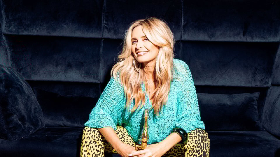 Candy Dulfer sitzt auf einem dunklen Sofa, hält ein Saxophon in der Hand und trägt ein türkisfarbenes Oberteil sowie gemusterte Hose, vor dunklem Hintergrund.