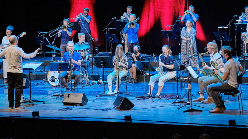Big Band mit Saxophonen, Posaunen und Trompeten während einer Aufführung auf beleuchteter Konzertbühne.