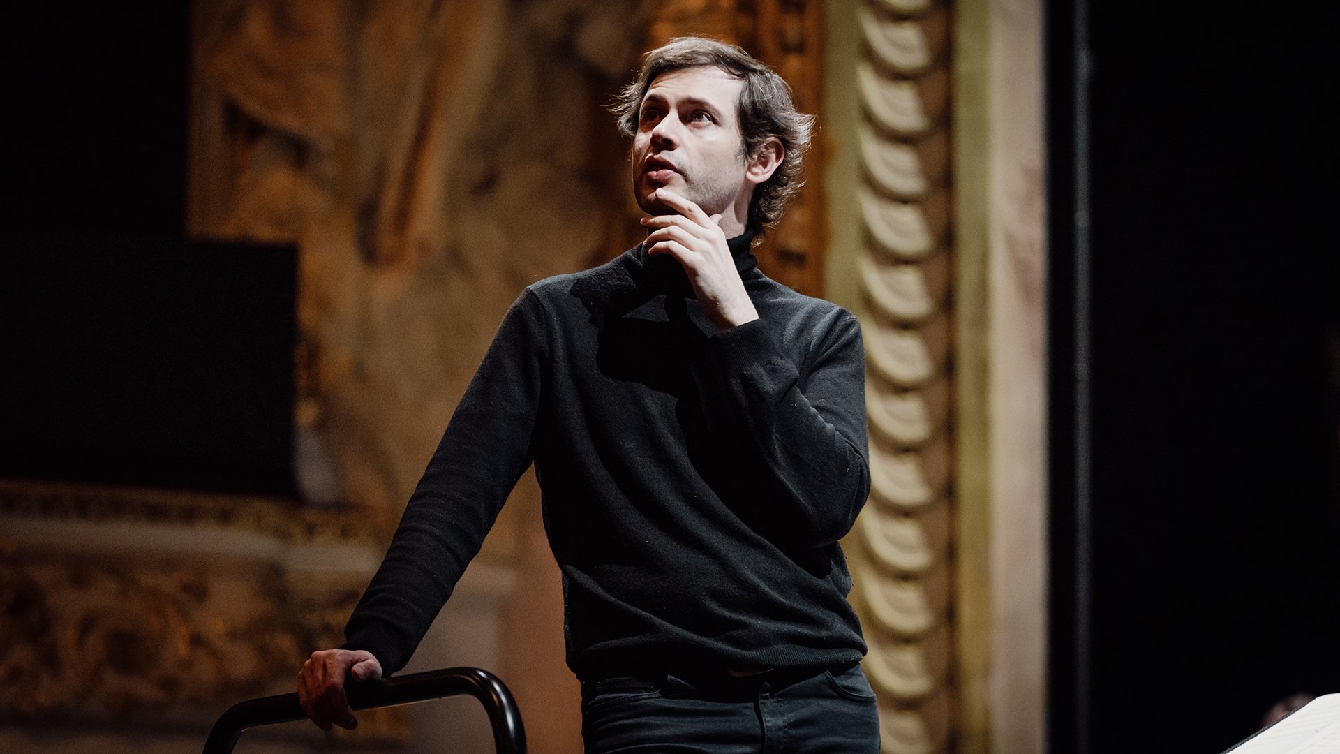 Maxime Pascal, an ein Dirigentenpult gelehnt, in dunkler Kleidung, vor dem Hintergrund eines kunstvoll verzierten Konzertsaals mit warmen Farbtönen und klassischen architektonischen Details.