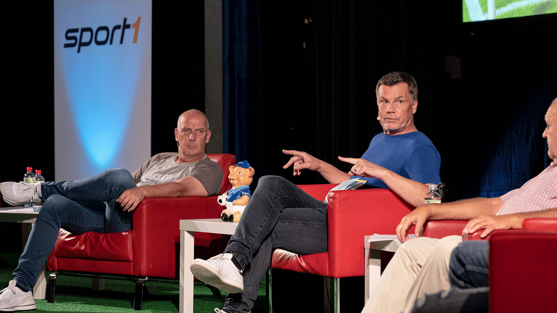 Podiumsdiskussion auf einer Bühne mit roten Sesseln. Mehrere Personen sitzen in Gesprächsrunden zusammen, während eine Person im blauen Shirt auf einen Gesprächspartner zeigt. Im Hintergrund ist ein Sport1 Logo zu sehen.
