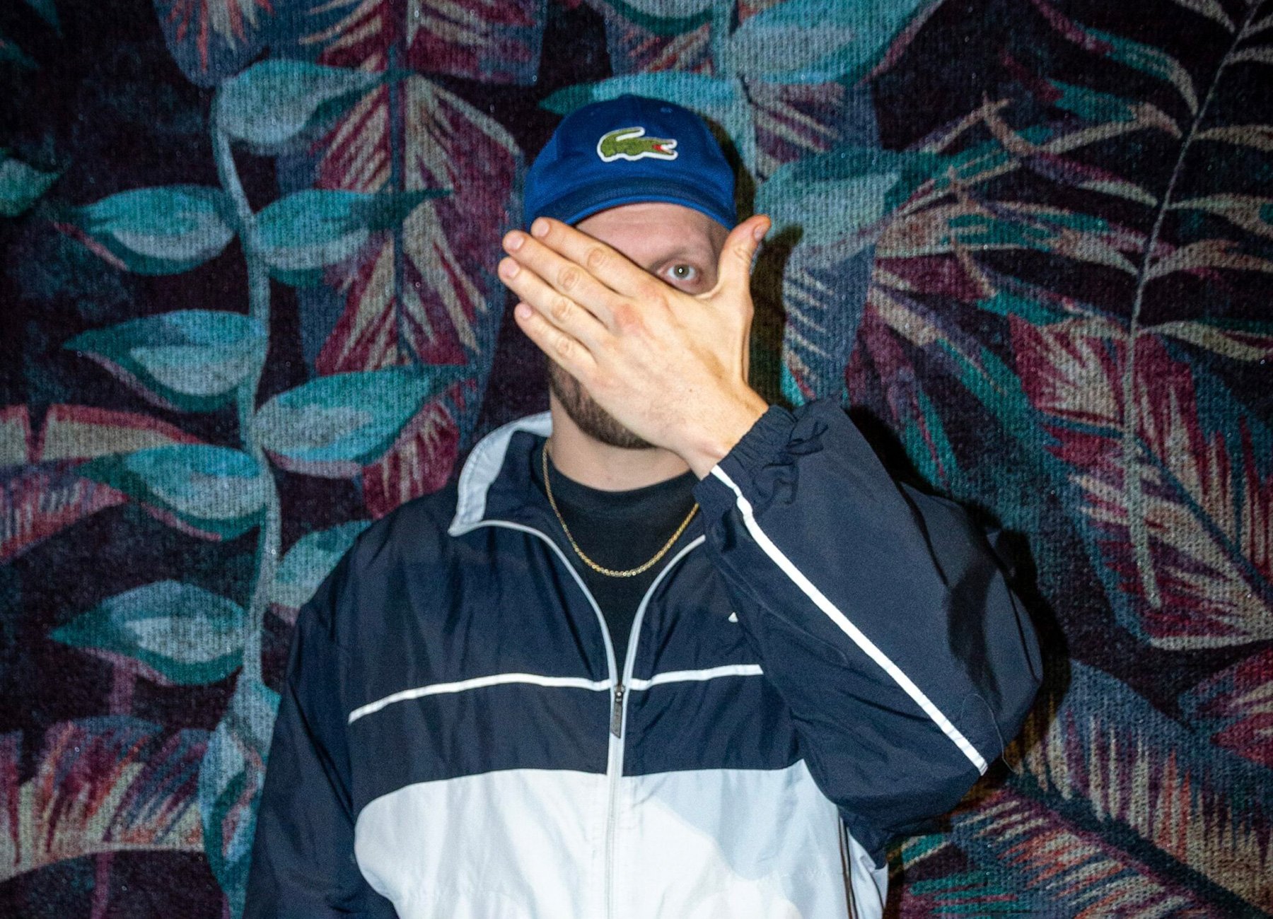 Mann mit blauer Kappe und blau-weisser Kleidung vor gemustertem Hintergrund, hält sich die Hand vor das Gesicht