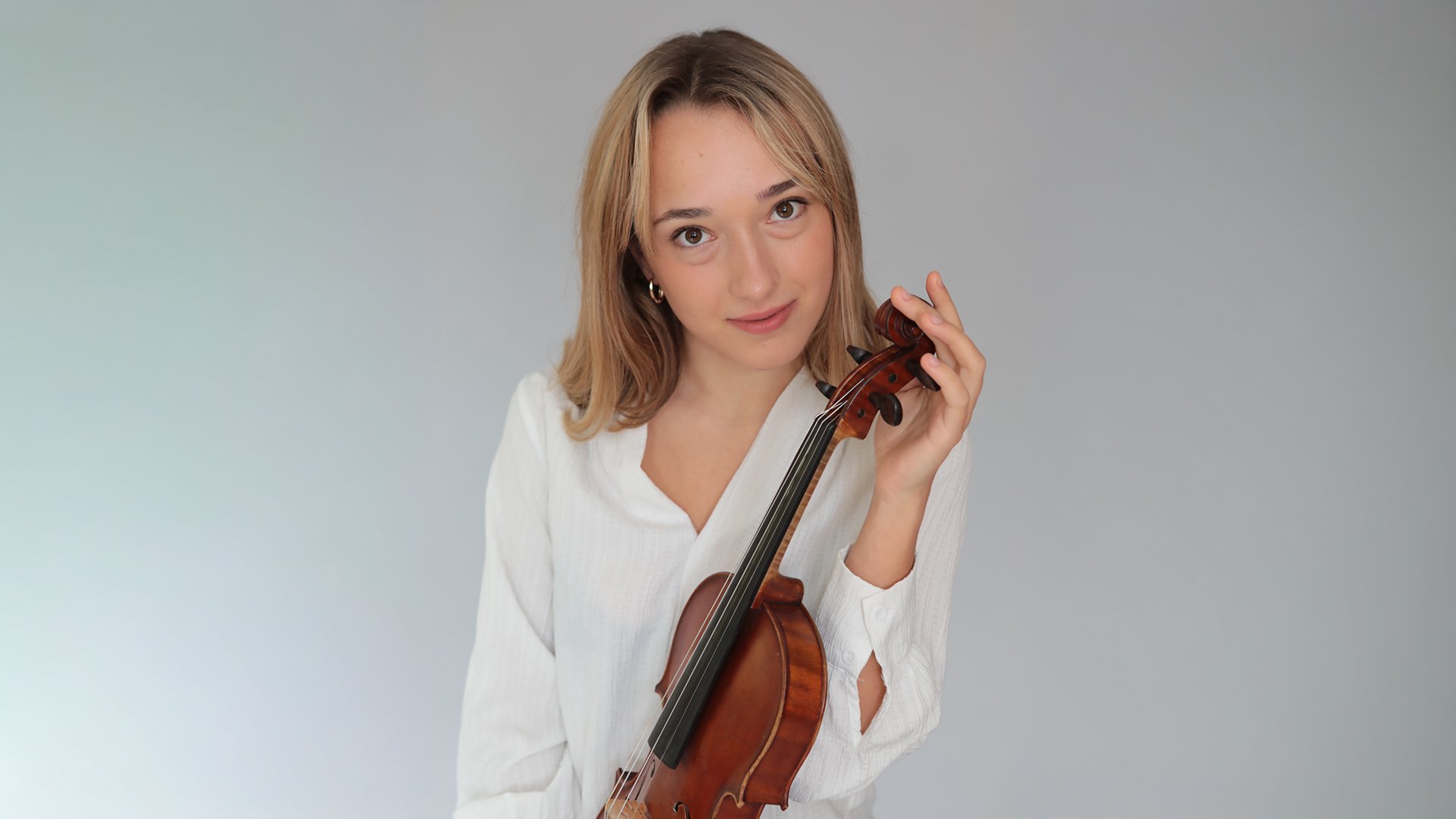 Porträtmotiv der Violinistin Bernadette Pihusch: Eine Person steht vor einem großen Fenster mit winterlicher Außenansicht und hält eine Violine nah am Oberkörper. Das Instrument wird mit einer Hand am Hals gefasst, die andere Hand liegt auf dem Korpus der Violine. Die Person trägt ein ärmelloses, dunkelrotes Konzertkleid; dezente Innenraumbeleuchtung und natürliche Lichtreflexe betonen Holzstruktur und Oberfläche des Instruments. Ruhige, warme Bildstimmung; Promofoto für ein klassisches Violinkonzert.