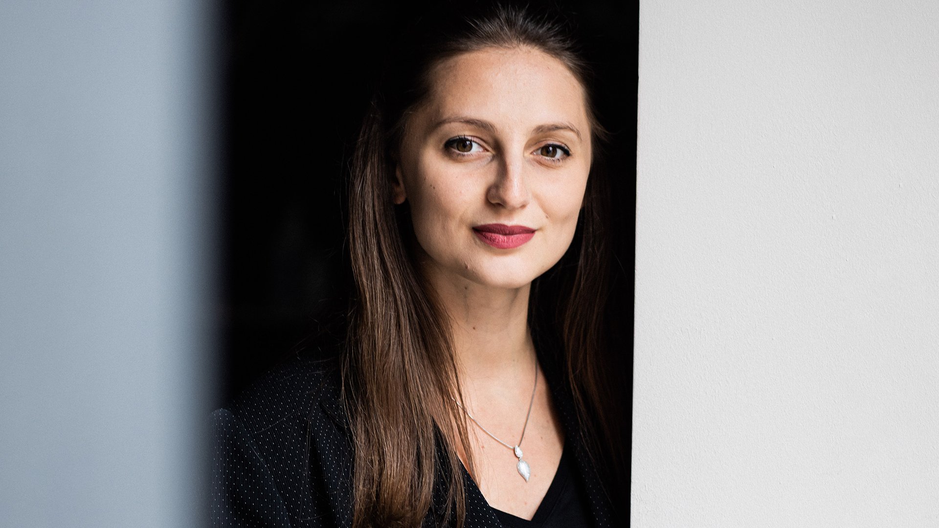 Die Künstlerin Rebeka Stojkoska lächelnd in schwarzer Kleidung vor dunklem Hintergrund, die Ränder des Bildes links und rechts sind hellblau und hellgrau