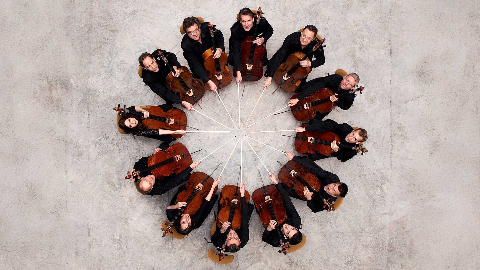 Promobild für das Event ‚Die 12 Cellisten der Berliner Philharmoniker‘: Zwölf Personen sitzen in einem Kreis auf Stühlen, halten jeweils ein Cello und richten die Bögen zur Mitte hin aus. Die Aufnahme zeigt die Gruppe aus der Vogelperspektive auf hellem Boden