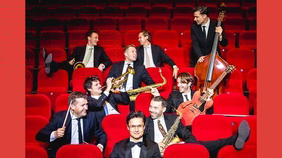 Foto für die Veranstaltung "DENNIS VAN AARSSEN & die DVA Bigband", mit den Künstlern auf roten Sesseln sitzend.