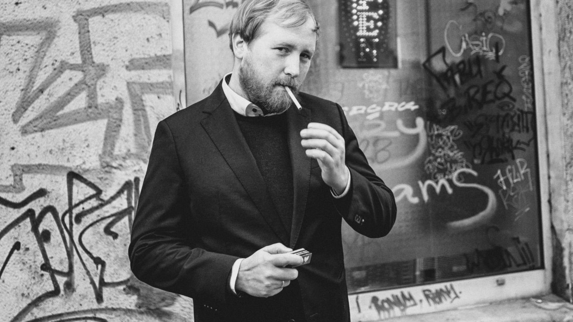 Julius Fischer in einem dunklen Anzug mit Zigarette im Mund steht vor einer mit Graffiti bedeckten Wand 